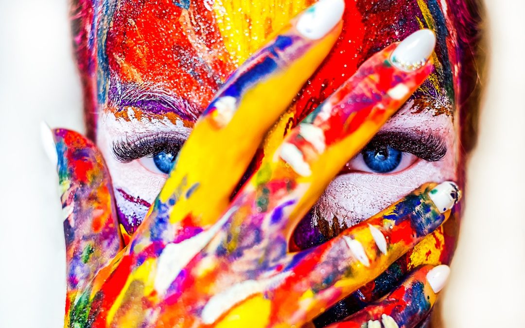 Colors painted on a woman’s face with eyes peering out between her fingers