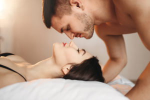 Man and woman in bedroom, woman lying face up on bed and man face down, nose to nose from opposite directions